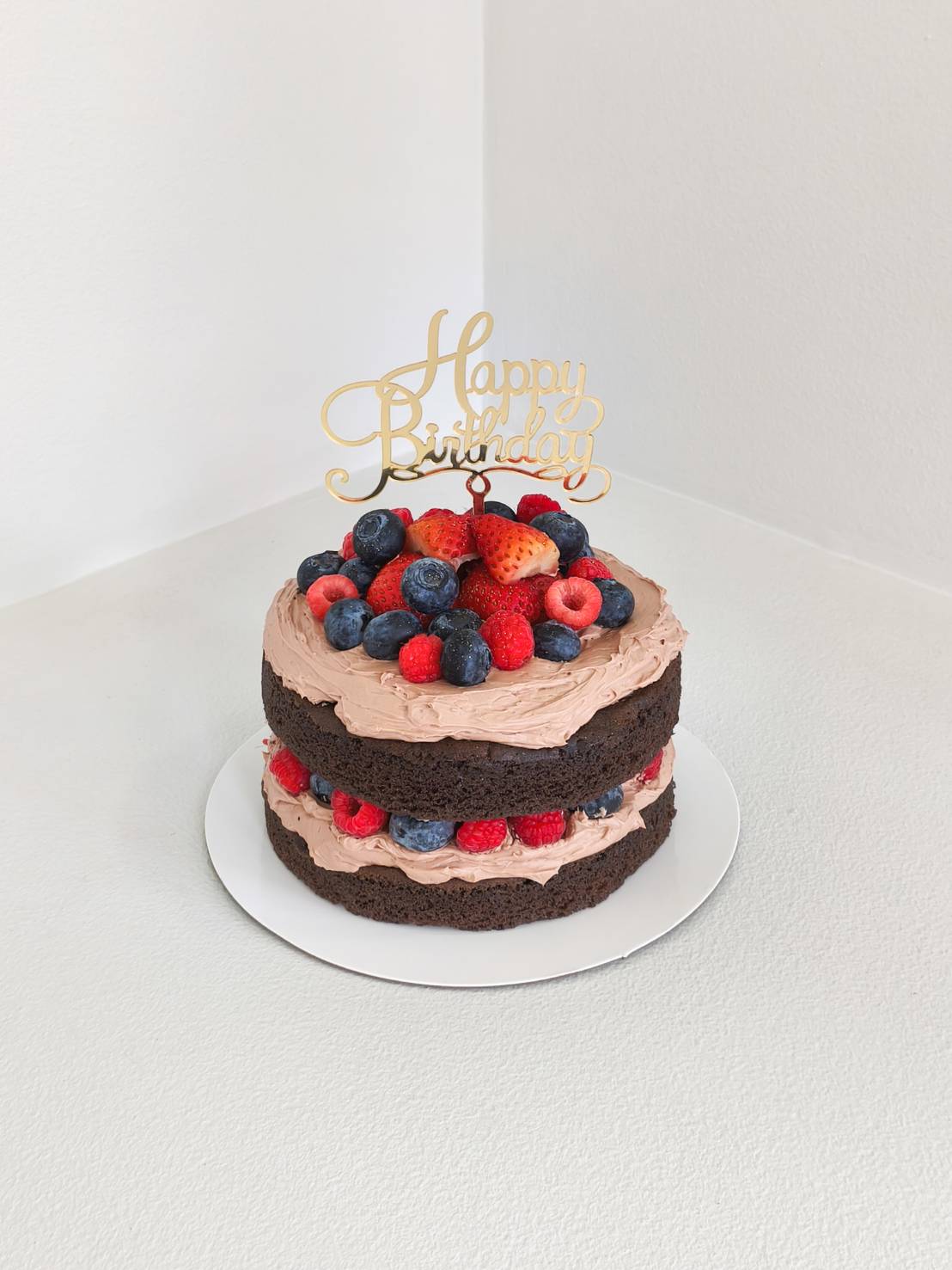Chocolate naked cake with layers of chocolate cream, topped and filled with fresh strawberries, blueberries, raspberries, and a gold “Happy Birthday” topper.