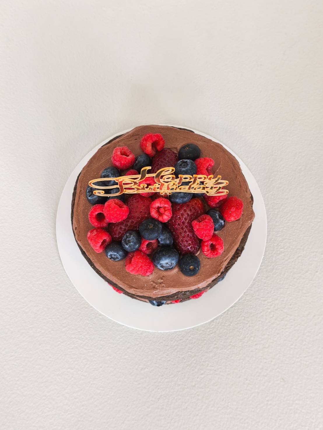 Chocolate birthday cake topped with fresh strawberries, blueberries, raspberries, and a gold “Happy Birthday” topper.