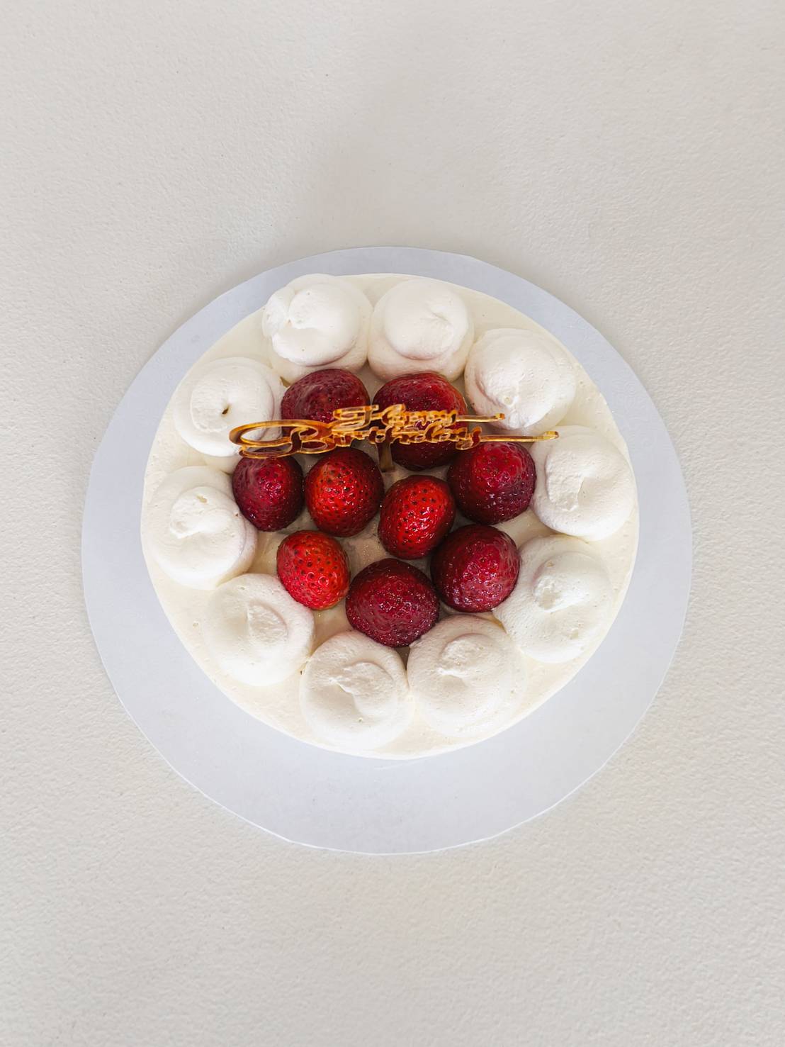 Top view of a fresh cream strawberry cake decorated with whipped cream swirls and whole strawberries, finished with a golden Happy Birthday topper.