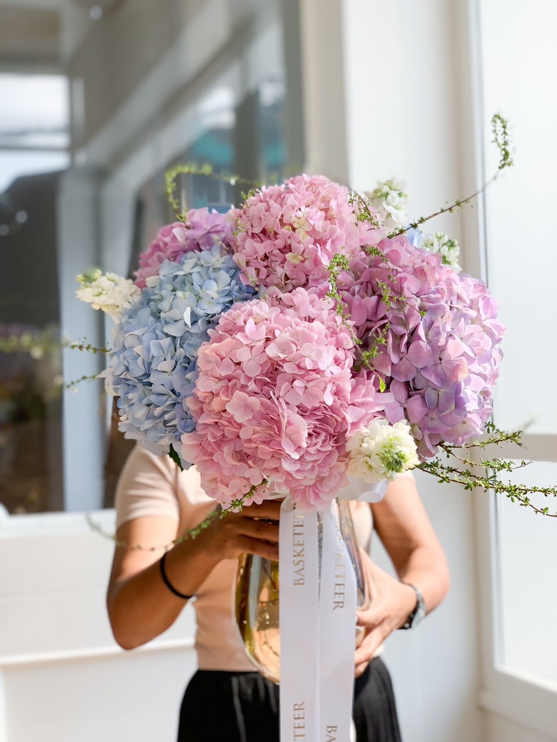 A person is holding a large bouquet of hydrangeas in varying shades of pink, blue, and purple. The bouquet exudes big hydrangea elegance and is tied with a white ribbon that has 