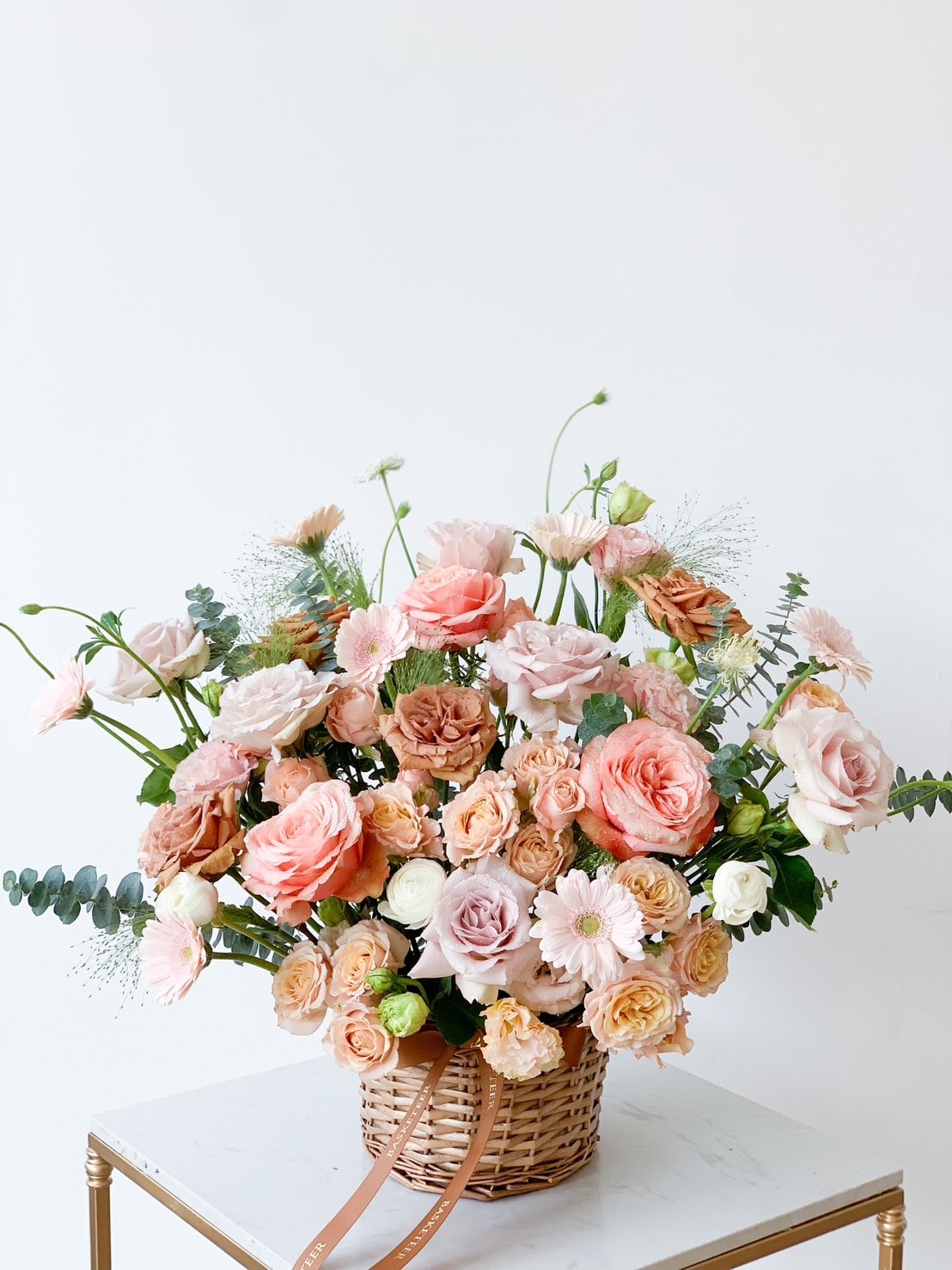 A wicker basket full of various roses and greenery sits on a white marble surface. The arrangement includes pink, peach, and white roses along with some delicate foliage, set against a plain white background. A brown ribbon is tied around the basket's handle.