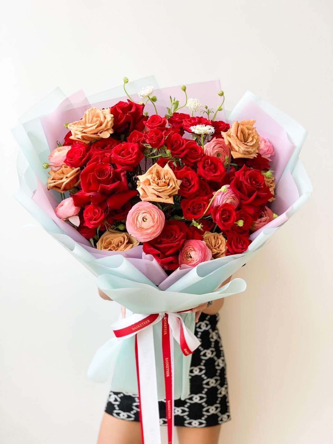 A person holding a large, beautifully arranged bouquet of roses. The bouquet contains red, pink, and peach roses wrapped in pastel-colored paper and tied with white and red ribbons. The background is plain white.