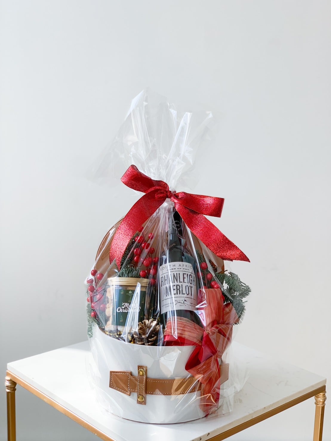A festive gift basket wrapped in clear cellophane and adorned with a large red bow. Inside, there is a bottle of wine, a small can, pinecones, and greenery. The basket has a holiday theme, resembling Santa's outfit with a white exterior, brown belt, and gold buckle.
