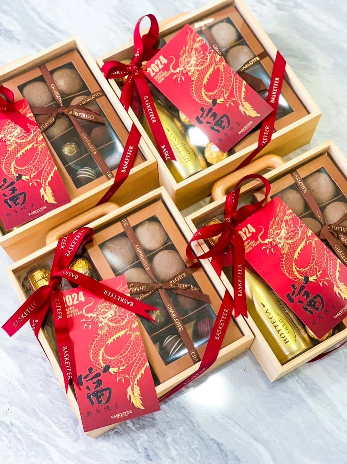 A set of four wooden gift boxes tied with red ribbons, displaying an assortment of chocolates and a golden bottle. Each box has a red card adorned with a gold dragon design and the year "2024". The boxes are placed on a white marble surface.