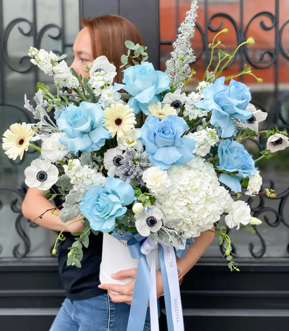 In a scene that exudes opulence and charm, a person with long graceful brown hair, partially visible, tenderly cradles an exquisite floral arrangement. This luxurious bouquet showcases elegant blue roses and pristine white hydrangeas, harmoniously blended with other delicate white and blue blooms amidst lush greenery. The arrangement is tastefully adorned with satin light blue and white ribbons. Prominently featured is a chic ribbon emblazoned with the Basketeer logo, meticulously tied to add an extra touch of elegance to this stunning ensemble designed for discerning tastes.