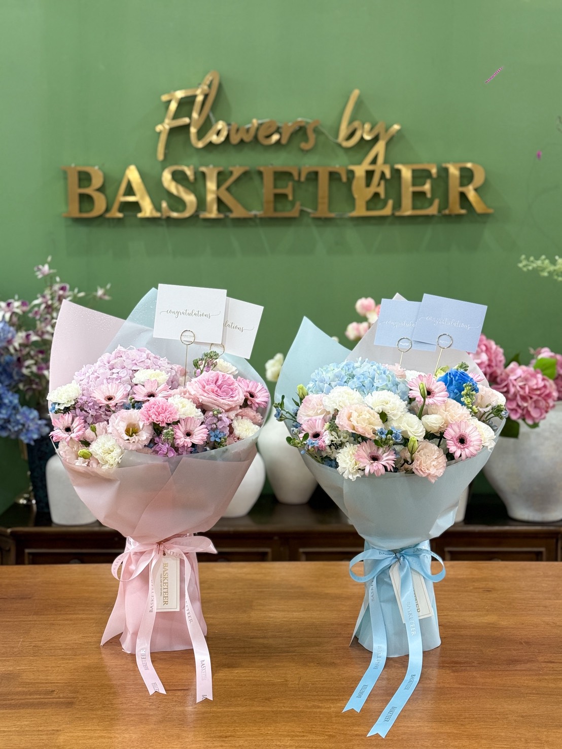 Two pastel bouquets, including the Vibrant Blue Lustre Bouquet with pink and blue wrapping, sit on a wooden table before a green wall with the gold 