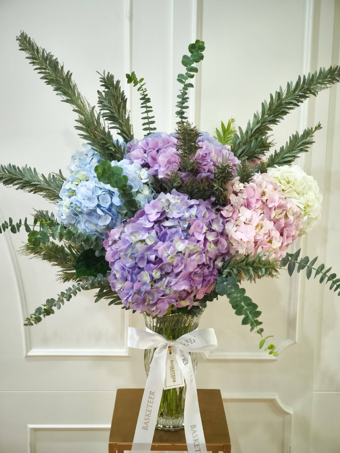 Elegant glass vase with pastel hydrangeas in shades of blue, purple, pink, and white, accented with lush greenery.