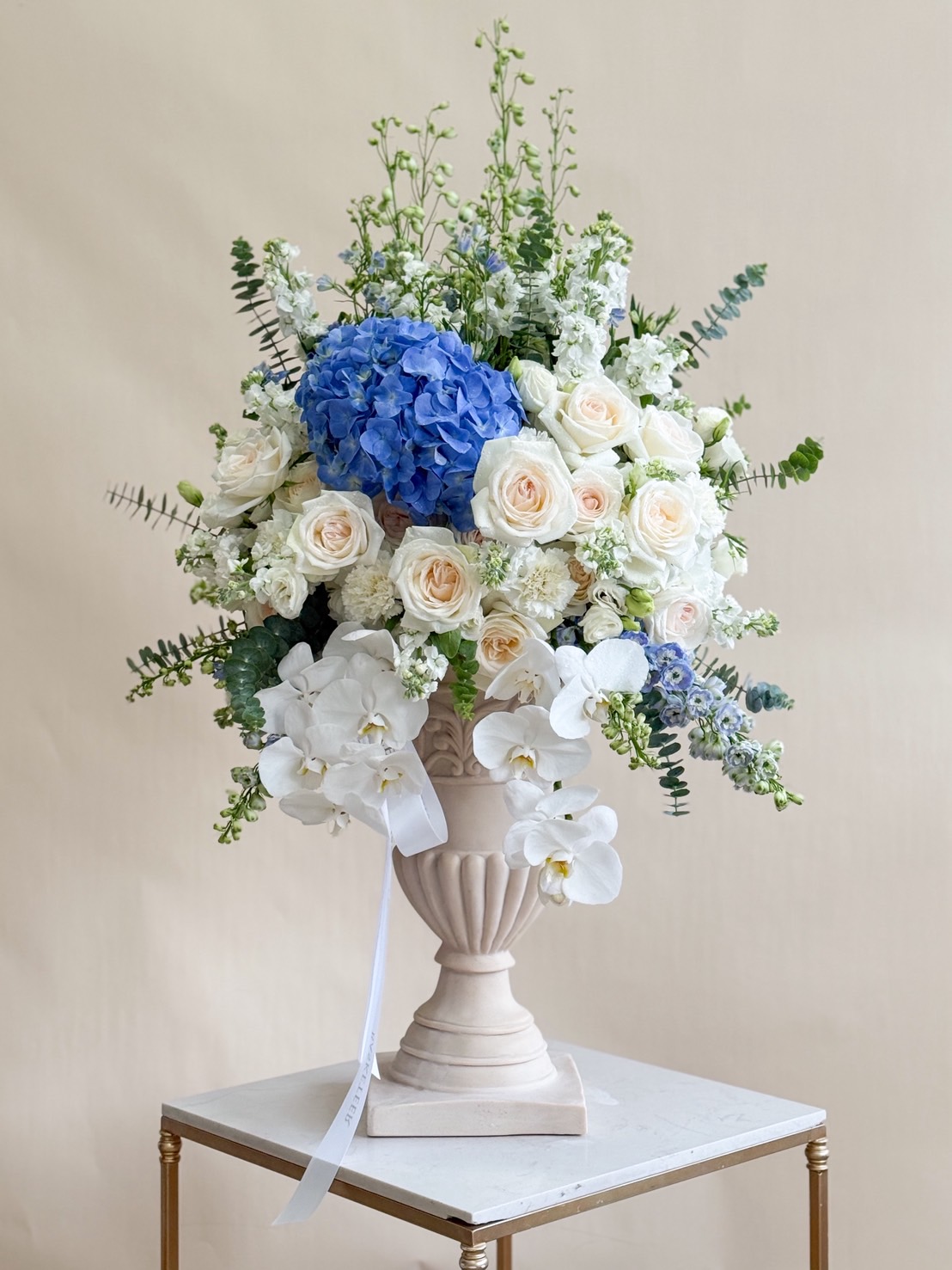 Tall pedestal vase with blue hydrangea, white orchids, ivory roses, and lush greenery arranged by Basketeer