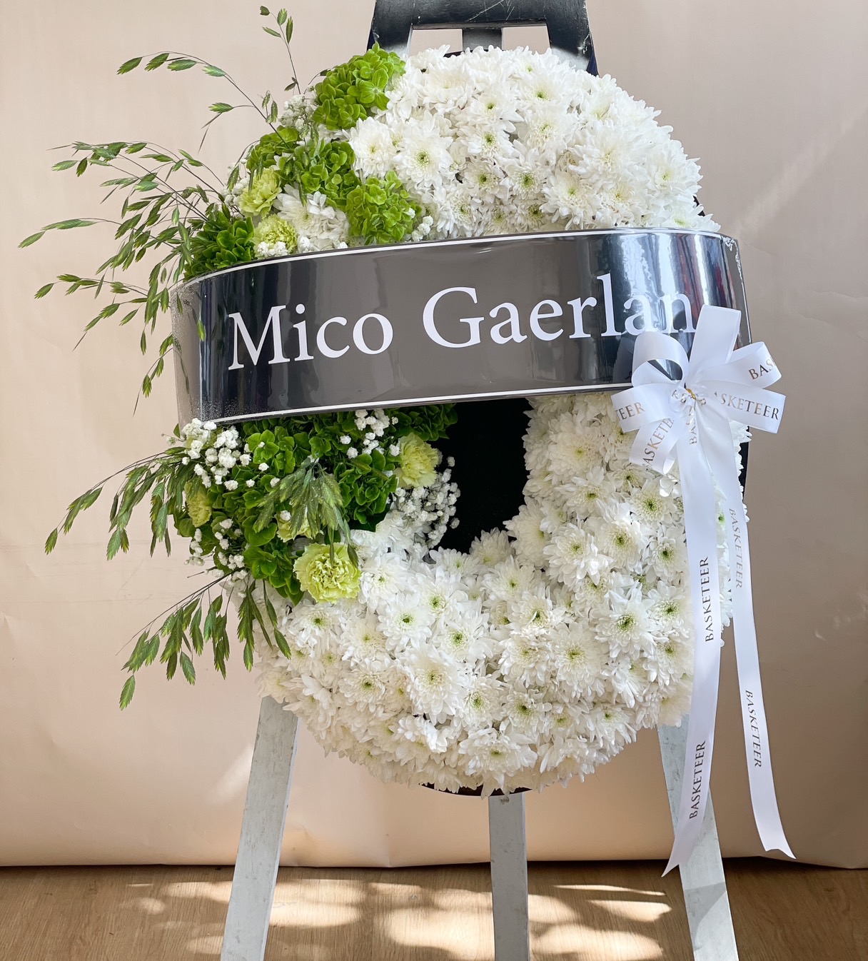 Elegant white floral wreath with chrysanthemums, greenery accents, and a custom name ribbon, displayed on a wooden stand.