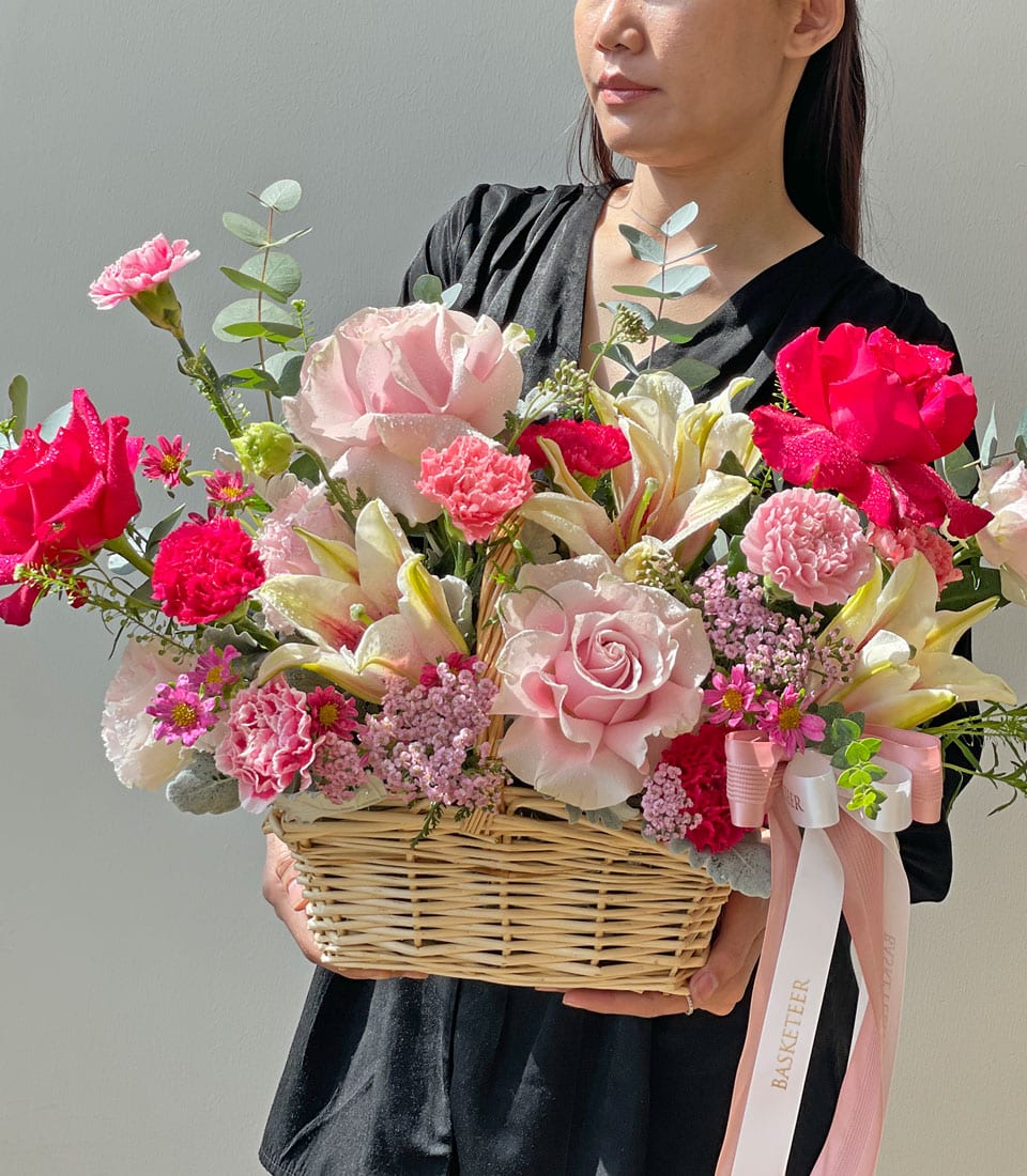 A person elegantly dressed in black showcases a luxurious wicker basket brimming with an enchanting array of vibrant flowers. This exquisite arrangement features breathtaking pink and red roses, delicate pink carnations, ethereal baby's breath, and lush eucalyptus leaves. Adorned with a sophisticated ribbon bearing the 