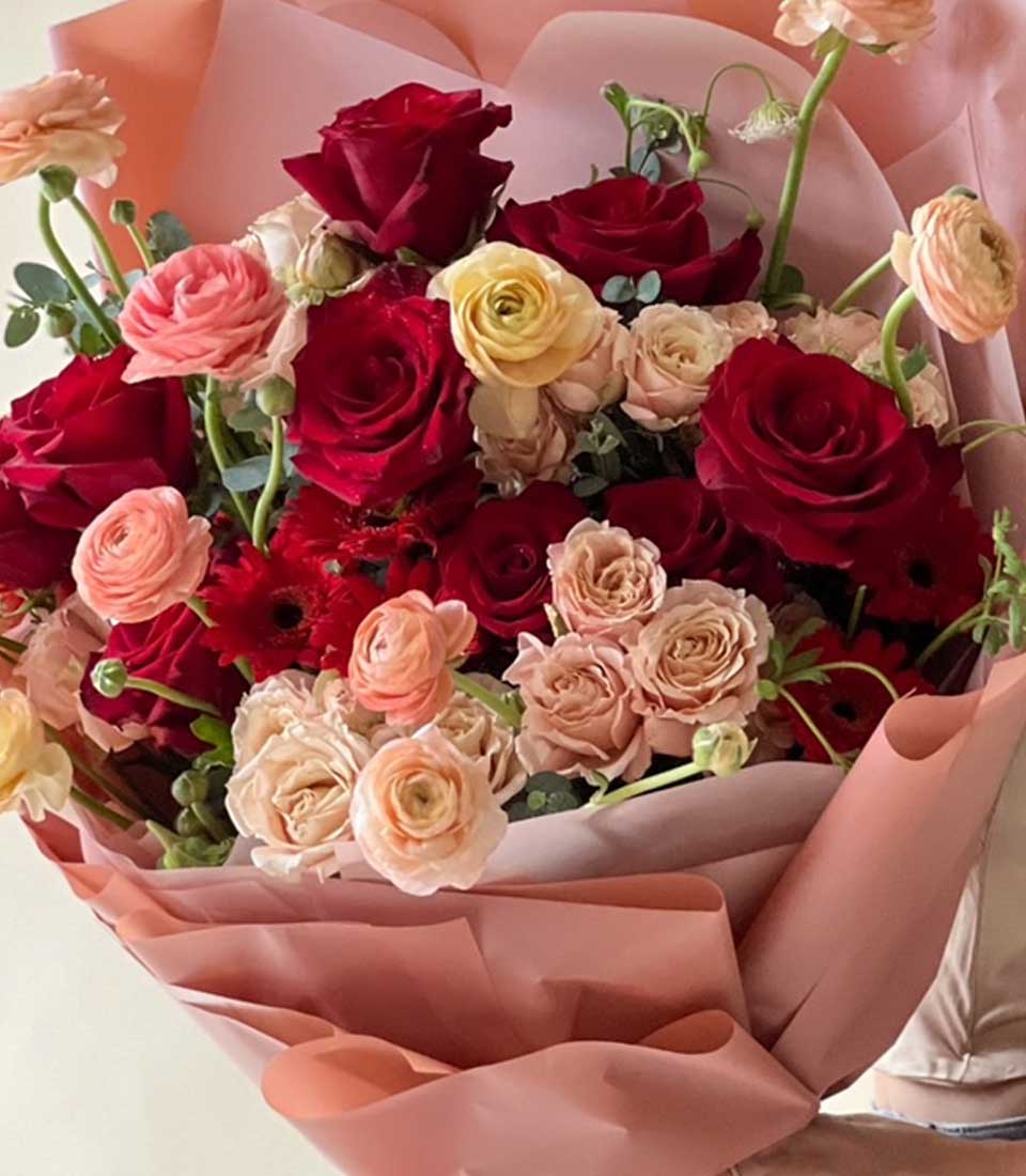An Opulent Crimson Rose Bouquet meticulously arranged with an exquisite selection of velvety red roses, delicate pale pink roses, and creamy white blooms. Encased in luxurious pink and peach wrapping paper, the bouquet radiates freshness and a vibrant charm. Nestled within this artful presentation is the iconic Basketeer ribbon, adding a touch of brand sophistication to an already elegant and romantic floral masterpiece.
