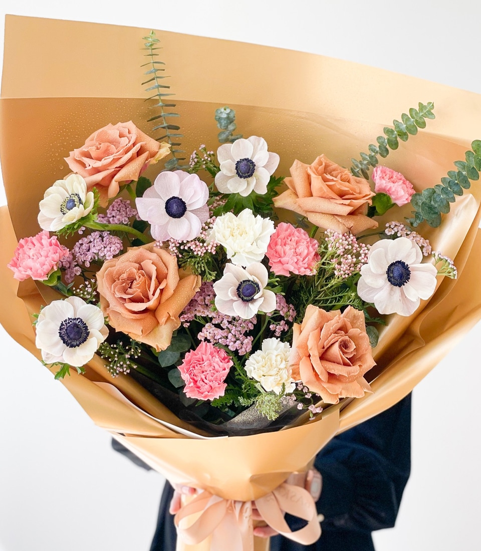 A Toffee Dreamland Anemone Bouquet wrapped in gold paper. The arrangement includes pale peach roses, white and pink anemones with dark centers, small pink carnations, and sprigs of eucalyptus leaves, held together with a light pink bow. The background is plain white.