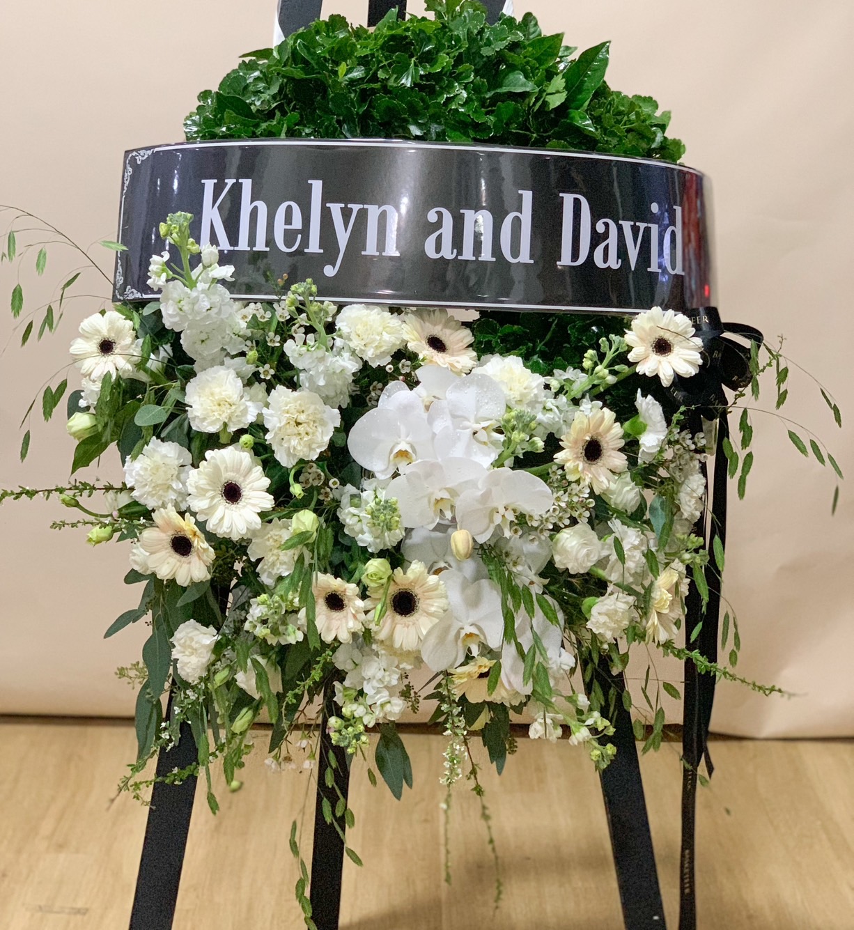 A delicate floral wreath, the Farewell Petals Wreath by Basketeer exudes luxury with its abundant green foliage and an artful arrangement of pristine white orchids, roses, and carnations gracing the bottom half. The elegant piece is thoughtfully presented on a sophisticated white easel, standing out against a simple gray backdrop. The scene captures a sense of inviting serenity and refined beauty.