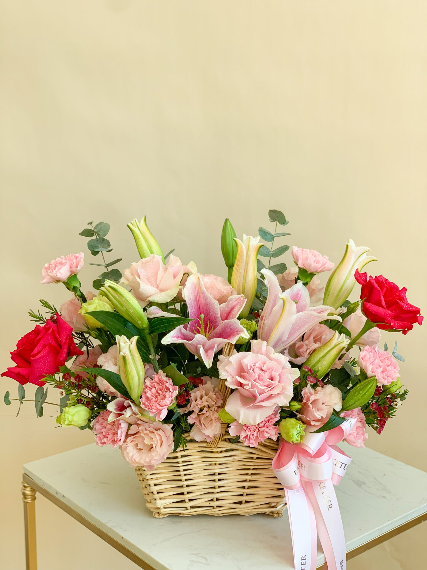 An exquisite wicker basket overflowing with an exquisite array of blooms, featuring elegant pink roses, pristine white lilies, vibrant red carnations, and delicate small pink flowers. Lush green leaves and fragrant eucalyptus sprigs intermingle beautifully within the arrangement. A luxurious pink ribbon elegantly tied around the basket proudly bears the 'BASKETEER' name. This stunning creation rests gracefully on a chic white table, creating an inviting ambiance that's both sophisticated and enchanting.