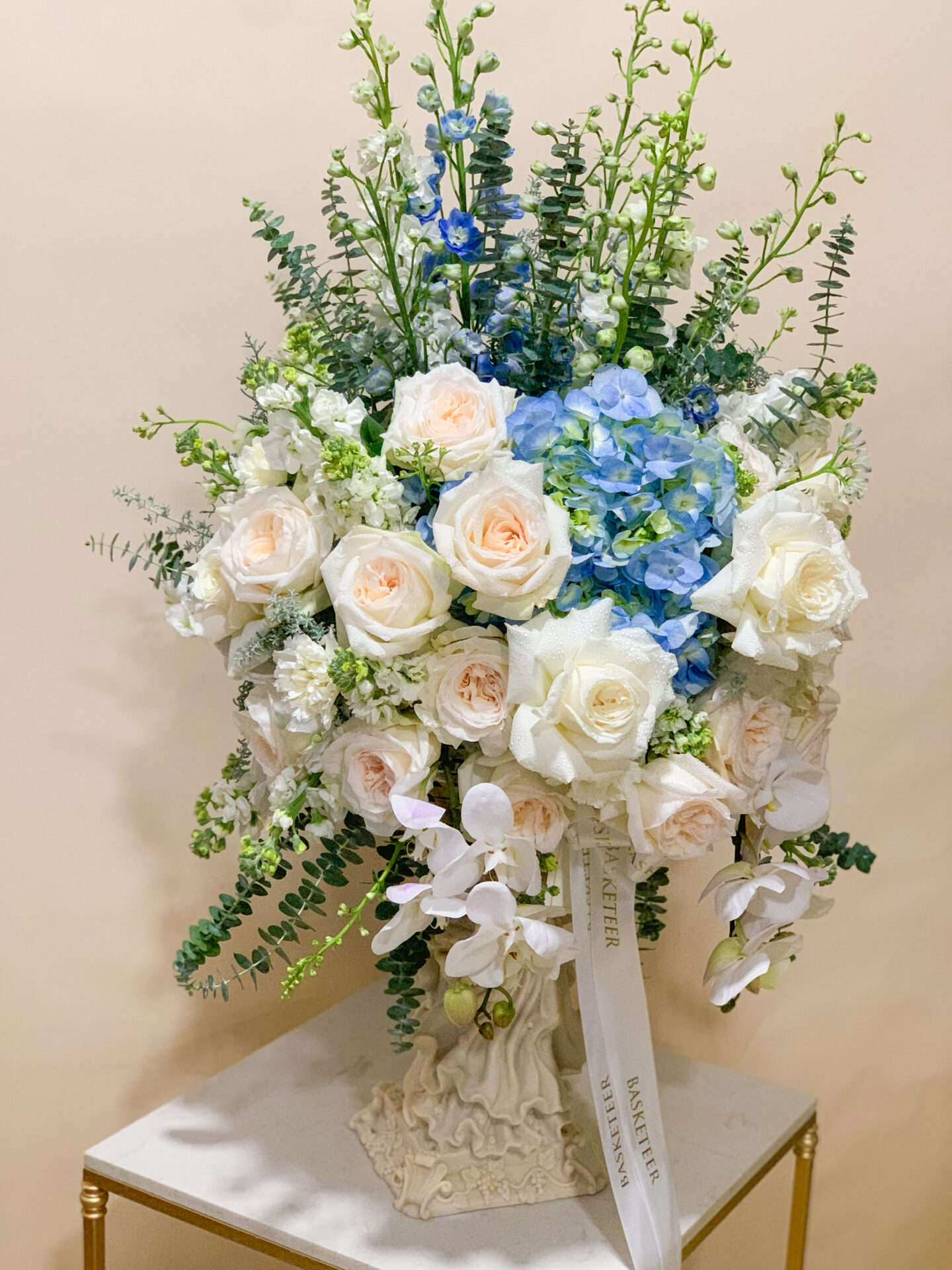 A person elegantly presenting a lavish bouquet of flowers. This exquisite arrangement boasts stunning white roses, delicate hydrangeas, and graceful orchids intertwined with lush green foliage, all nestled in a premium vase adorned with White Mondial Roses. Partially hidden by this breathtaking floral display, the individual stands gracefully against a backdrop of an opulent white paneled wall. A luxurious Basketeer ribbon subtly complements the scene, adding a touch of refinement and brand distinction.