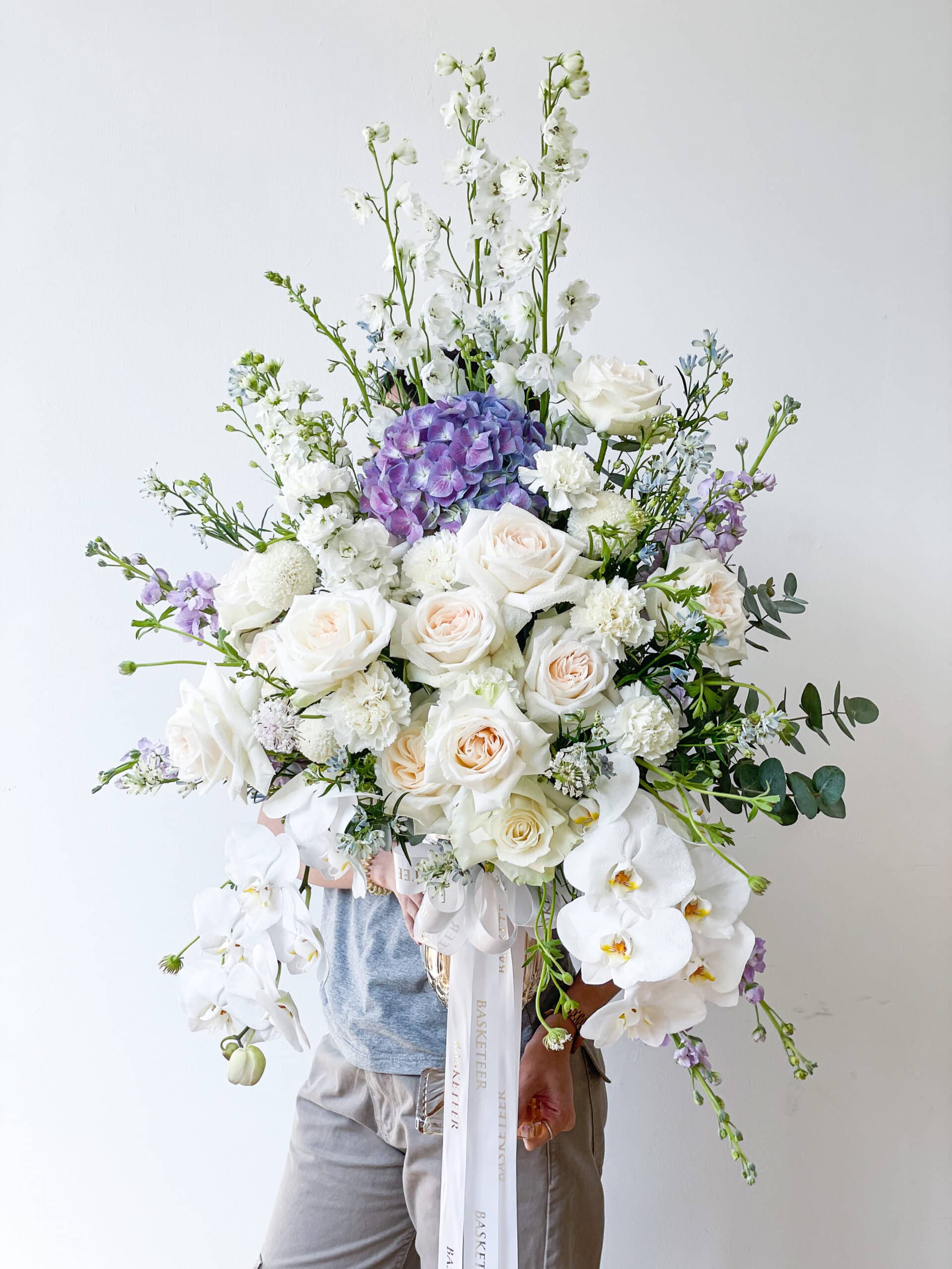 A person holding a large, elegant floral arrangement featuring white Mondial roses, orchids, and a cluster of purple hydrangeas in a premium vase against a plain white background. The bouquet also includes green foliage and delicate white and lavender flowers, secured with white ribbons.