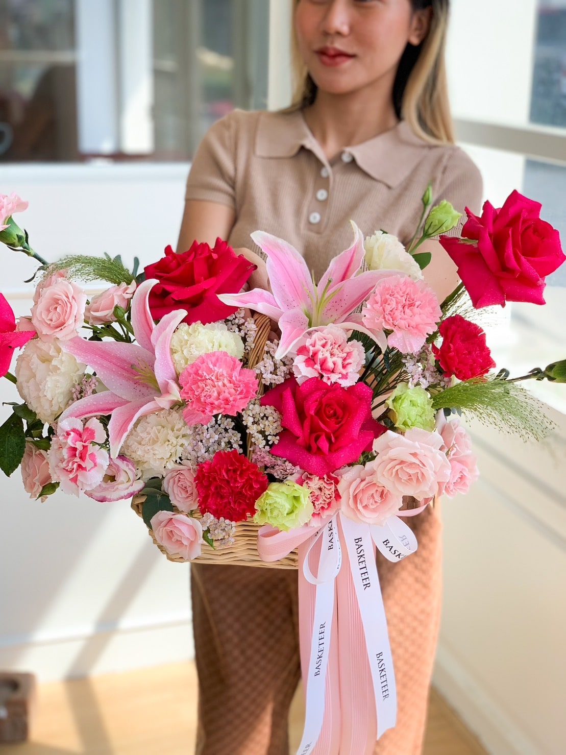 A woman in a beige top and checkered skirt holds a basket brimming with an enchanting floral arrangement. The bouquet features pink lilies, red and pink Mondial roses, white hydrangeas, and green carnations. The basket is adorned with white ribbons, creating a perfect harmony of colors.