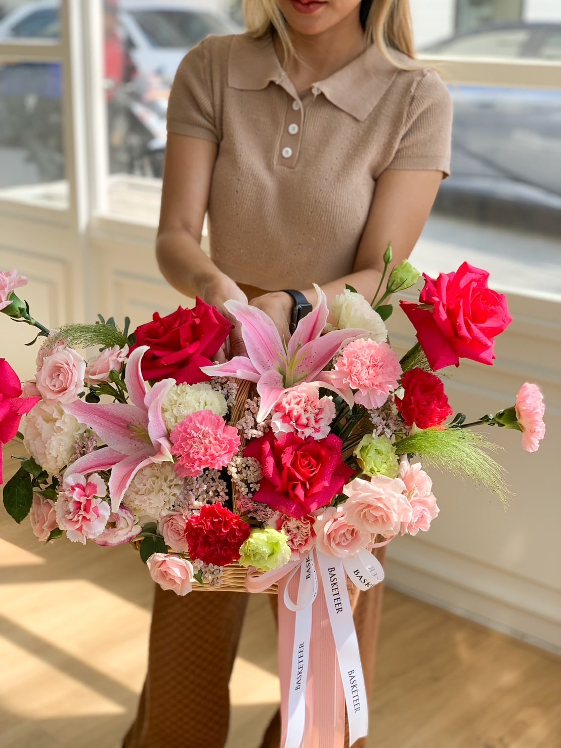 A woman with enchanting blonde hair, wearing a beige shirt, is holding a large floral arrangement. The bouquet includes vibrant pink and red roses, lilies, and carnations, tied with a ribbon. She is standing indoors near a window, radiating an aura of Mondial harmony.