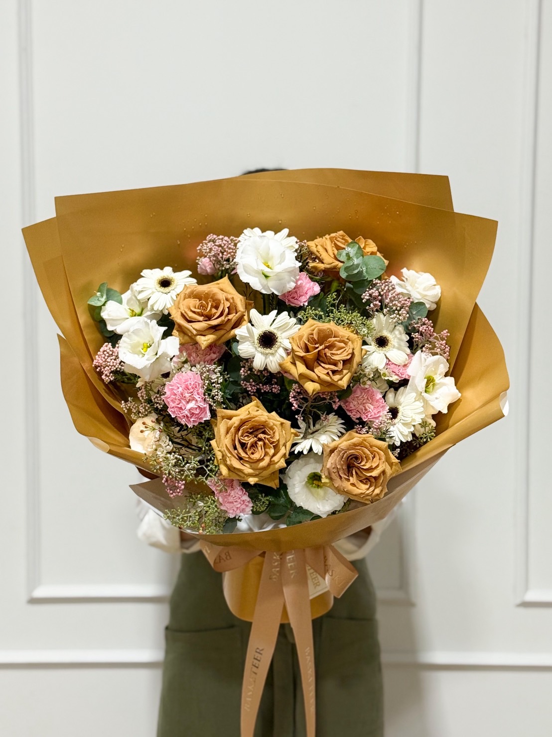 Luxury bouquet with golden toffee roses, white daisies, pink carnations, lisianthus, and eucalyptus wrapped in golden paper.