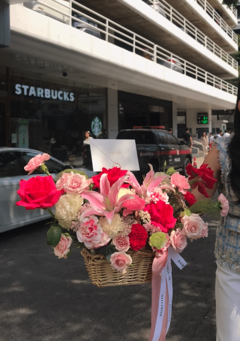 A person on the right holds a basket filled with an enchanting arrangement of flowers, including roses and Pink Mondial lilies, along with a white card. They stand on a street with a Starbucks, several parked cars, and apartment balconies visible in the background.