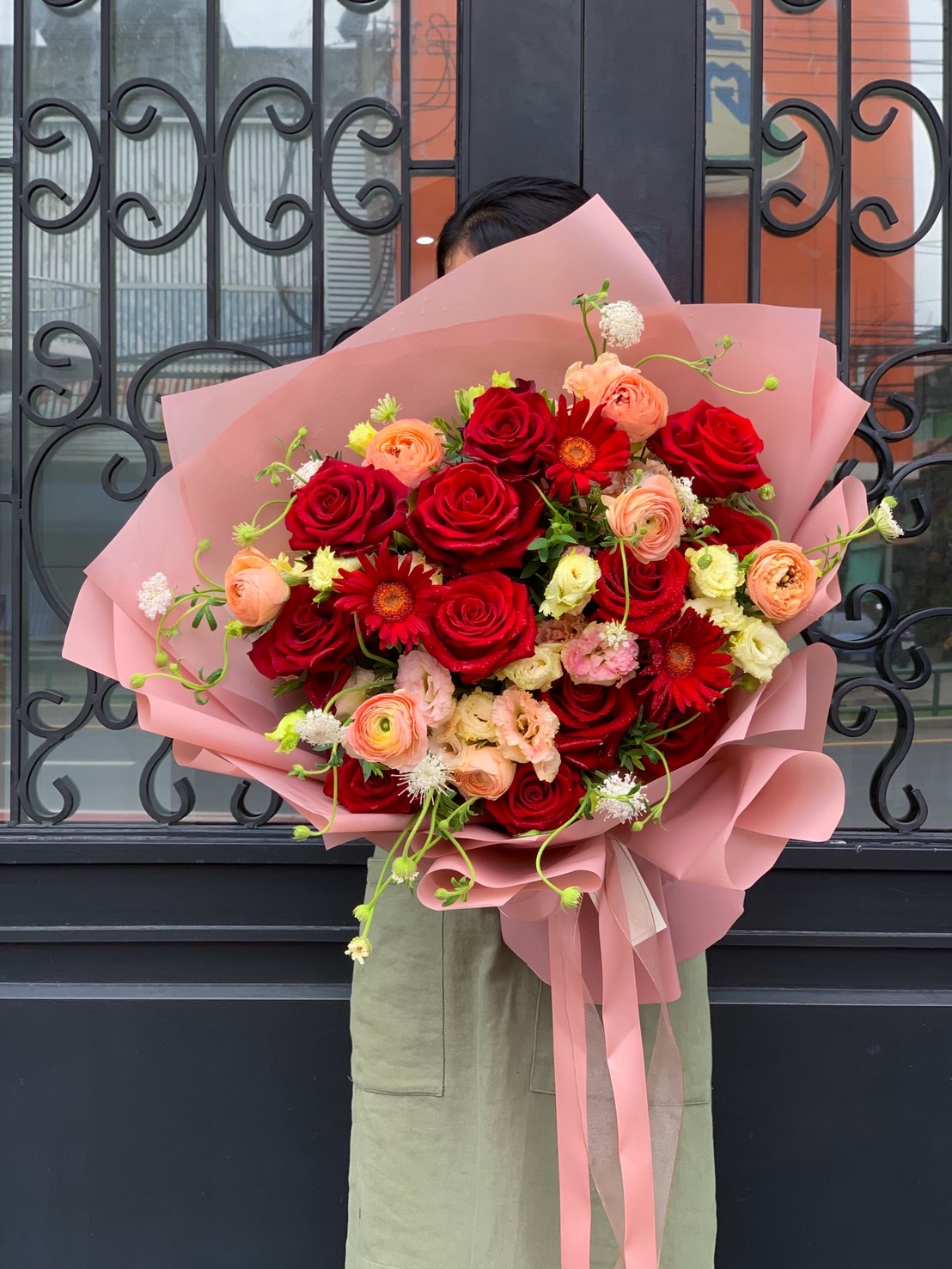 Large luxury bouquet with red roses, peach ranunculus, and red gerberas wrapped in pink paper, held in front of black iron doors.