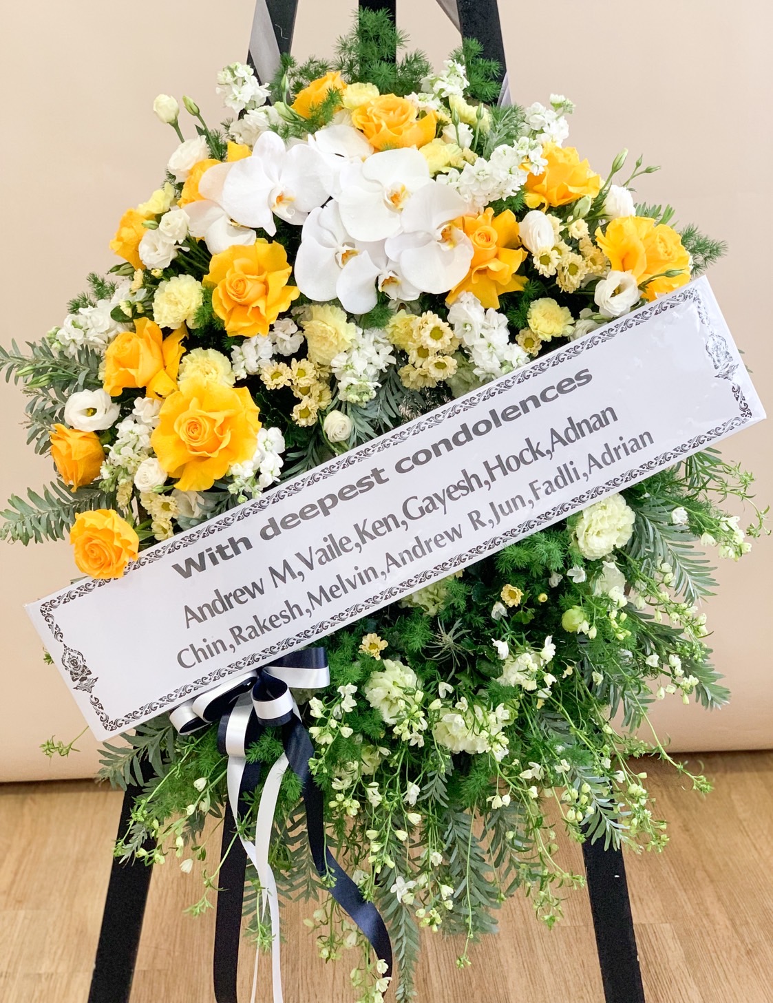 Condolence flower stand with yellow roses, white orchids, and soft greenery – sympathy tribute with message ribbon.