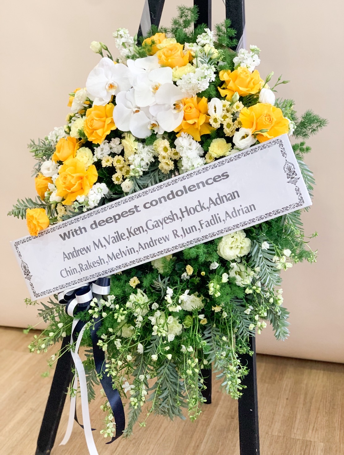 Condolence flower stand with yellow roses, white orchids, and soft greenery – sympathy tribute with message ribbon.
