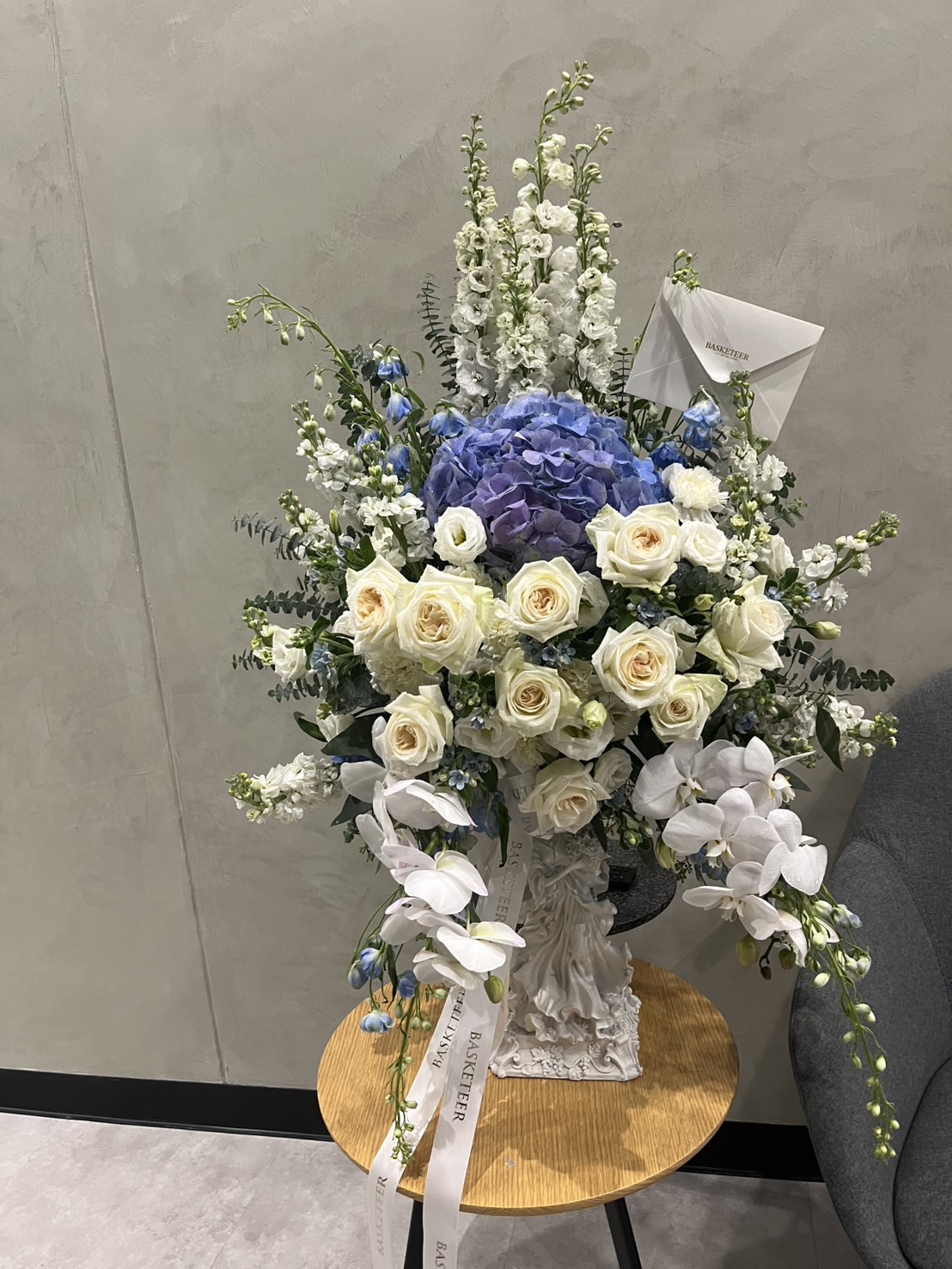 A tall floral arrangement in a premium vase with White Mondial Roses, white orchids, blue hydrangeas, and greenery sits on a small round wooden table. A card and white ribbons add an elegant touch to the bouquet.