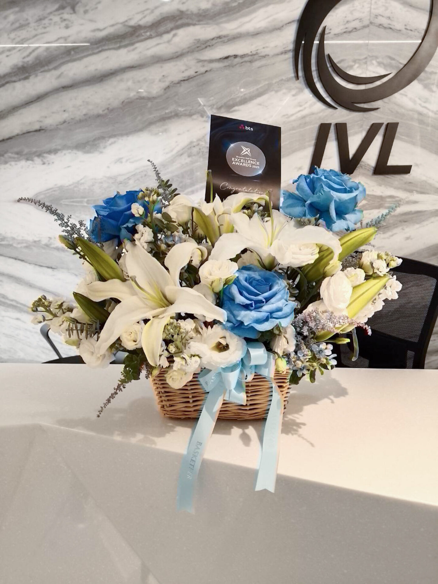 Elegant flower basket with blue roses, white lilies, lisianthus, and seasonal blooms, decorated with a light blue satin ribbon.