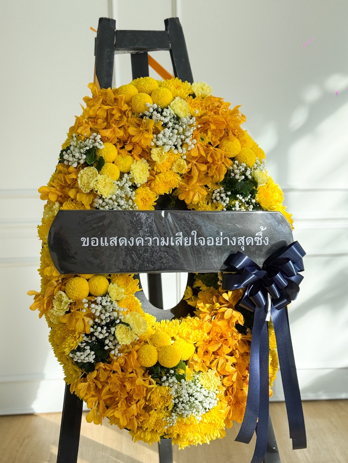 A Peaceful Yellow Flowers Wreath with white blooms and a navy blue ribbon is displayed on a stand. A Thai message on a black banner across the centre reads, 