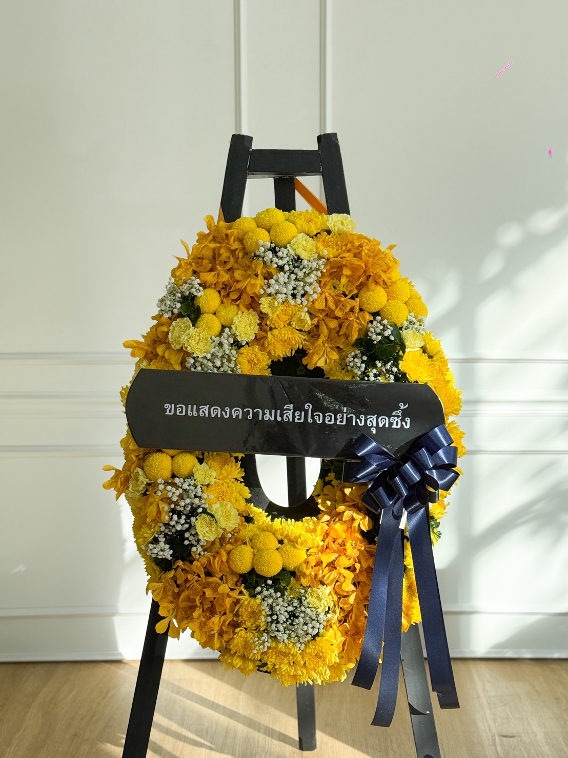 A Peaceful Yellow Flowers Wreath with a dark blue ribbon and a black sign bearing Thai text is displayed on a black stand against a white wall, bathed in gentle sunlight.