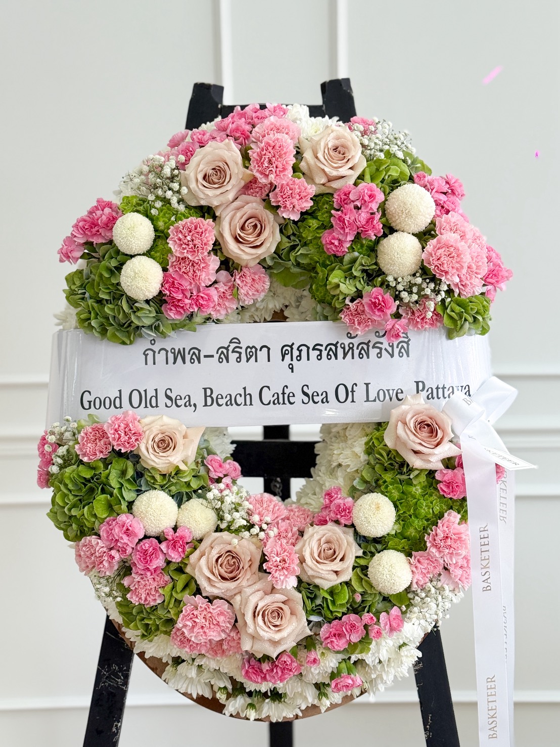 An Elegant Pastel Flowers Wreath featuring pink roses, white chrysanthemums, and lush green foliage is displayed on a stand. A ribbon with Thai text and the English words "Good Old Sea, Beach Café Sea Of Love, Pattaya" is attached.
