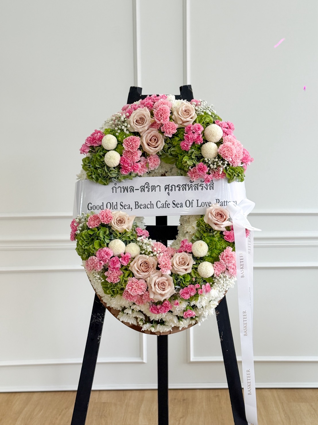 An Elegant Pastel Flowers Wreath with pink, white, and green blooms stands on a black easel. A white ribbon across the wreath reads, "Good Old Sea, Beach Cafe Sea Of Love Pattaya." The background is a white panelled wall.