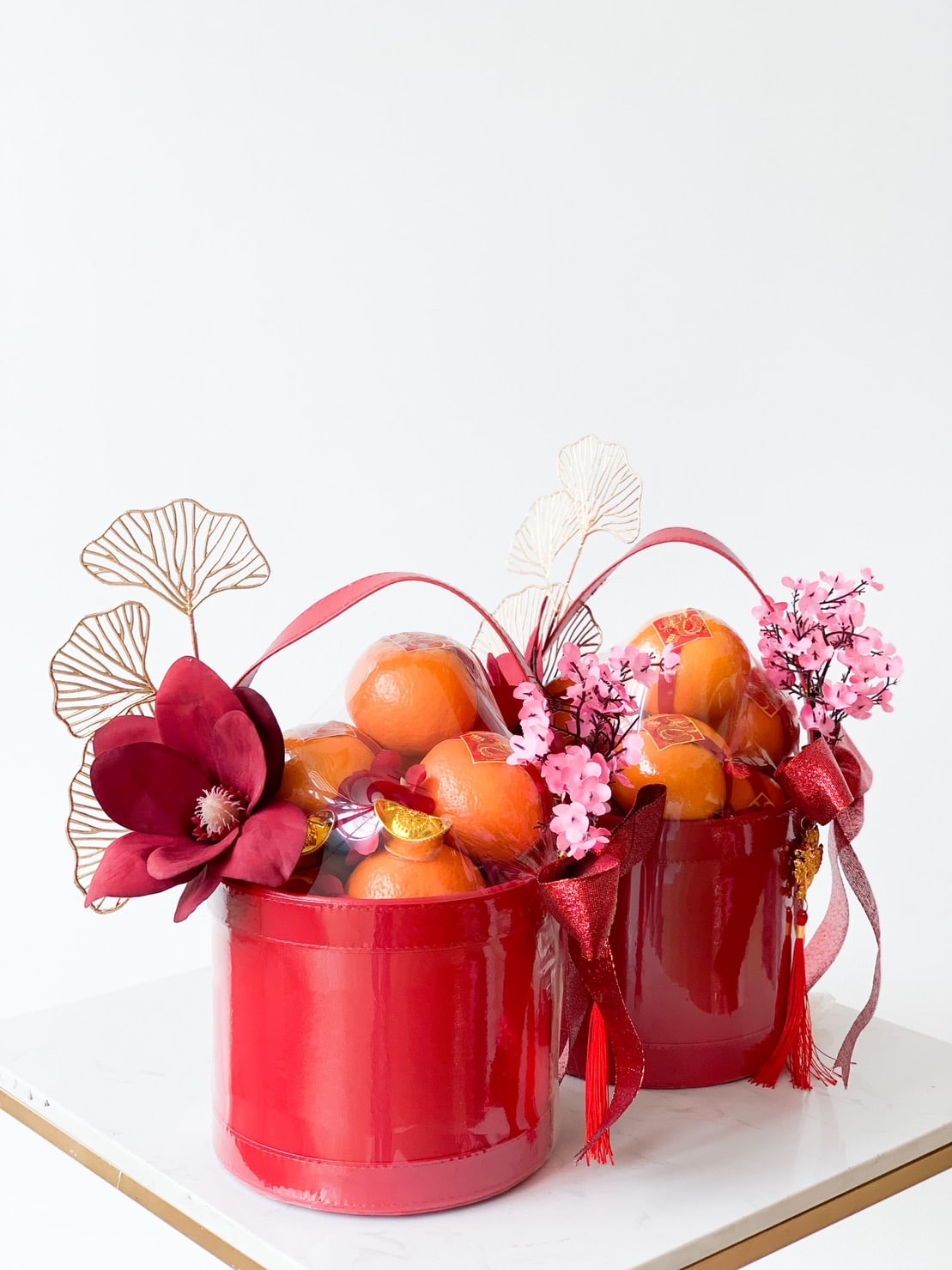 Two red baskets filled with oranges and decorated with vivid pink and red flowers, along with delicate gold accents. Each basket is tied with red ribbons and sits on a white surface. The background is plain white, emphasizing the vibrant colors of the decorations.