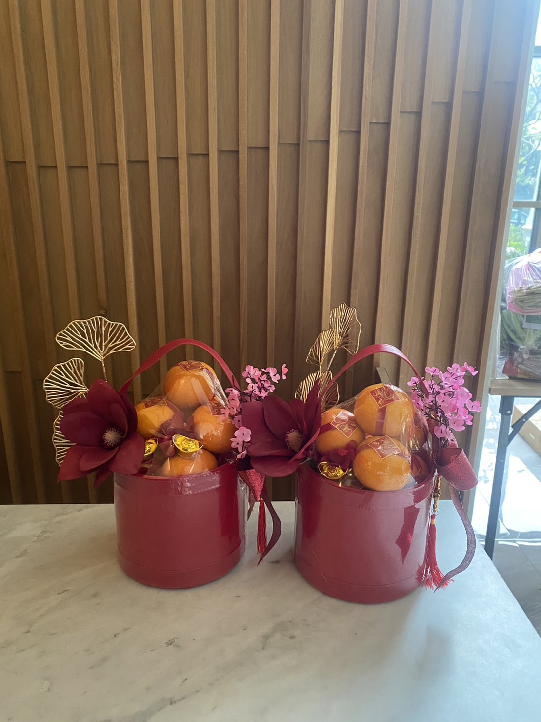 Two red buckets are filled with oranges, adorned with gold leaves, large red flowers, and pink blossoms. The buckets are placed on a marble surface with a wooden panel background. Small golden wrapped candies are scattered among the oranges.