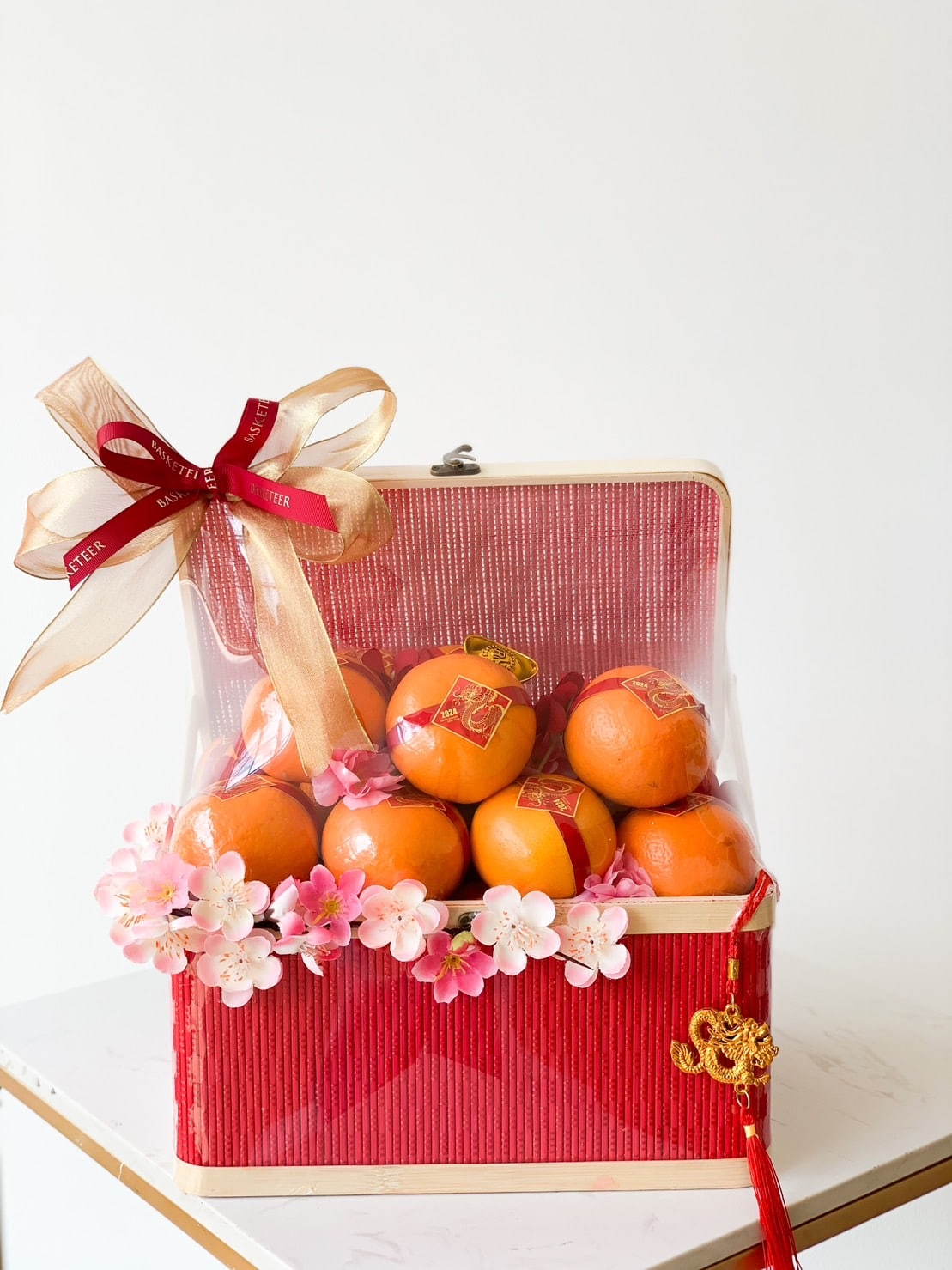 A red and gold gift box contains multiple oranges decorated with stickers, topped with a red and gold ribbon, and adorned with pink artificial flowers. The box is festive, possibly for a celebration like CNY, radiating harmony in Mandarin Delights Gift, with an elegant presentation.