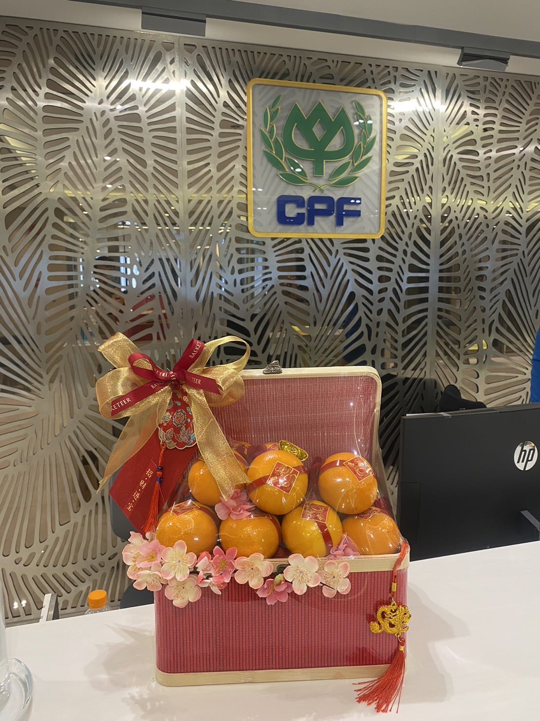 A festive basket filled with oranges, adorned with red and golden bows and pink flowers, sits on a white countertop. The background features a decorative panel with a CPF logo and a wall with foliage patterns. An HP computer monitor is partially visible, capturing the essence of CNY: Harmony in Mandarin Delights Gift.
