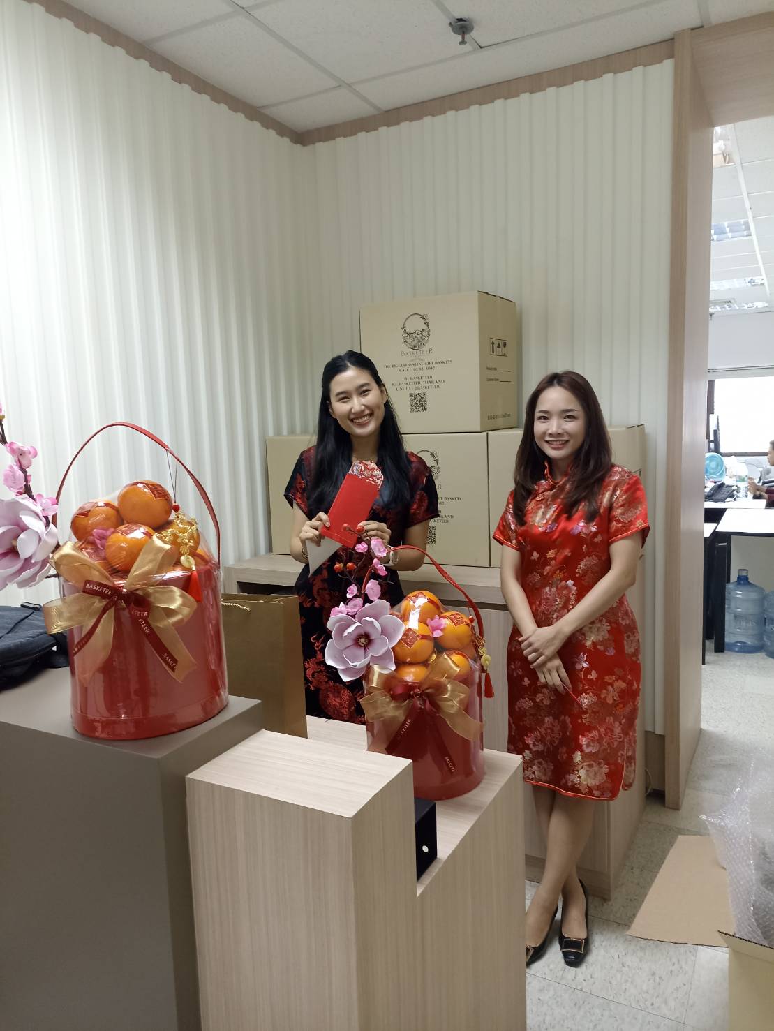 Two women wearing red Chinese traditional dresses stand in an office room, each holding a red envelope. Festive red containers with oranges and decorative flowers, including the sought-after Orange Bliss Box for CNY, adorn the table. In the background, cardboard boxes are stacked neatly.