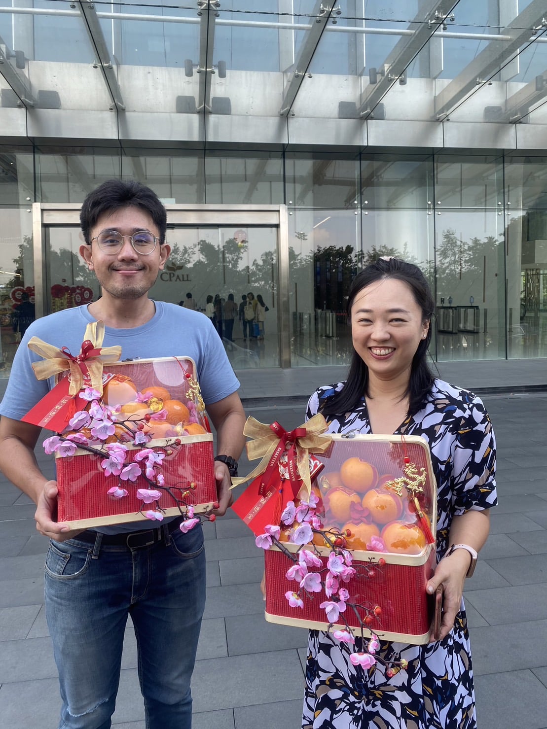A man and a woman stand outside a building, smiling and holding red baskets filled with oranges, decorated with flowers and ribbons. The man, in a grey t-shirt and jeans, and the woman, in a patterned dress, exude harmony in Mandarin Delights as they celebrate CNY.