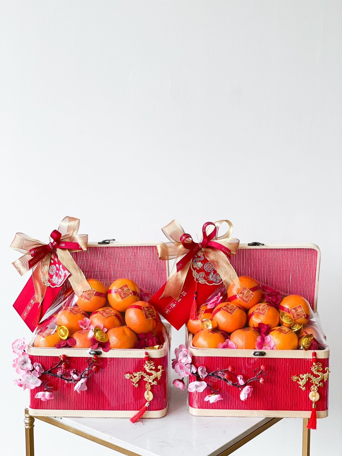 Two ornate red boxes, filled with fresh oranges and decorated with pink blossoms, golden bows, and red ribbons celebrate CNY. Embossed with intricate patterns and Chinese characters, these Harmony in Mandarin Delights gifts are displayed against a plain white background for a festive occasion.
