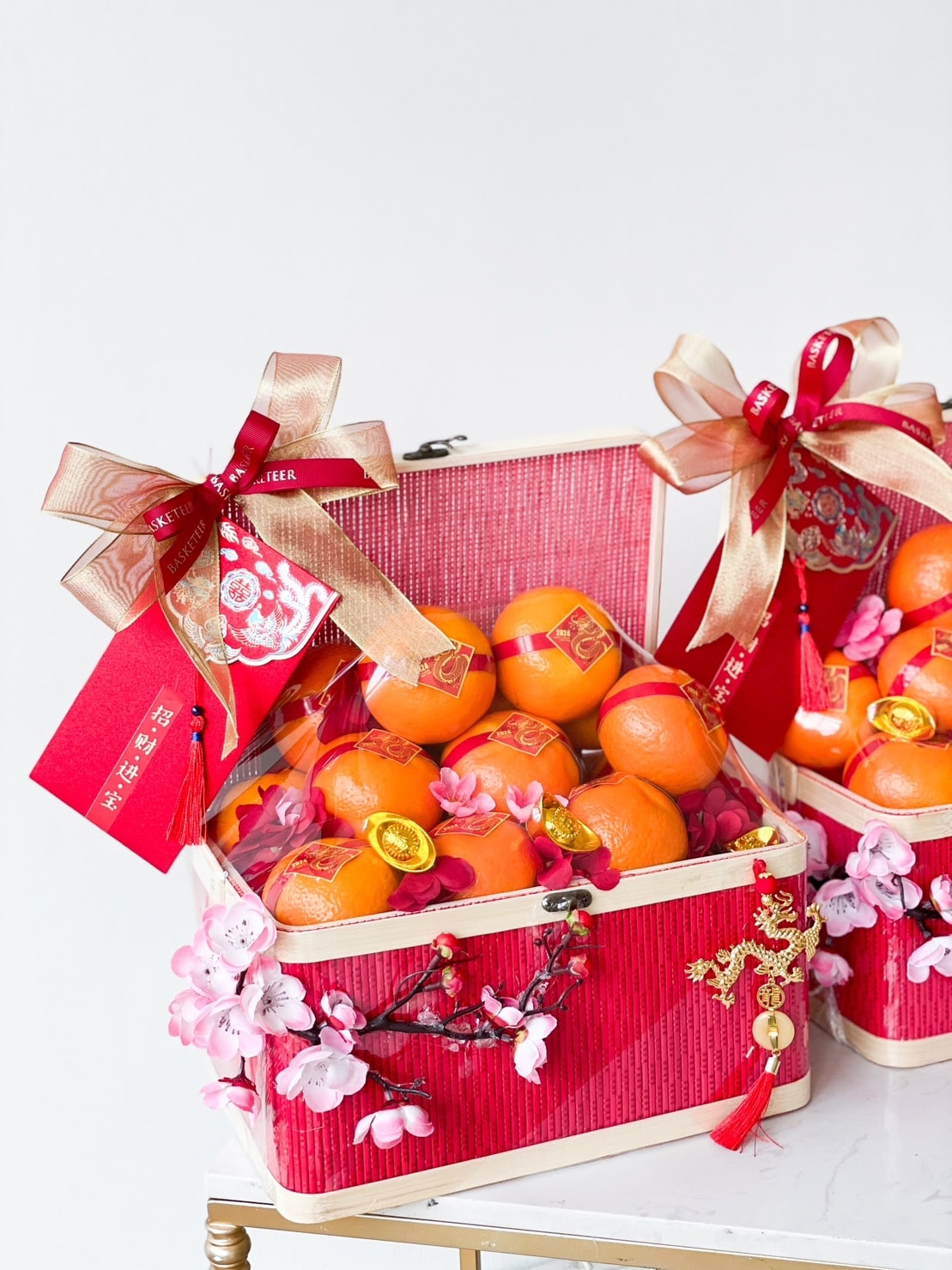 Two ornate gift boxes are filled with ripe oranges, gold ingots, and adorned with pink cherry blossoms. Each box, part of the Harmony in Mandarin Delights Gift for CNY, is decorated with red and gold bows and red envelopes, symbolizing prosperity and good fortune. The scene is bright and festive.