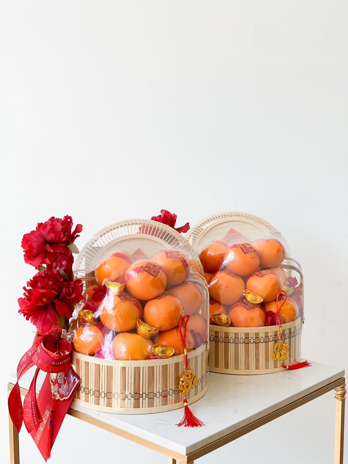 Two baskets, each filled with ripe oranges, are placed on a small table. The baskets, known as the Imperial Mandarin Elegance Basket, are decorated with red flowers, ribbons, and golden ornaments. The arrangement appears festive, possibly for a CNY celebration such as the Lunar New Year. The background is plain white.