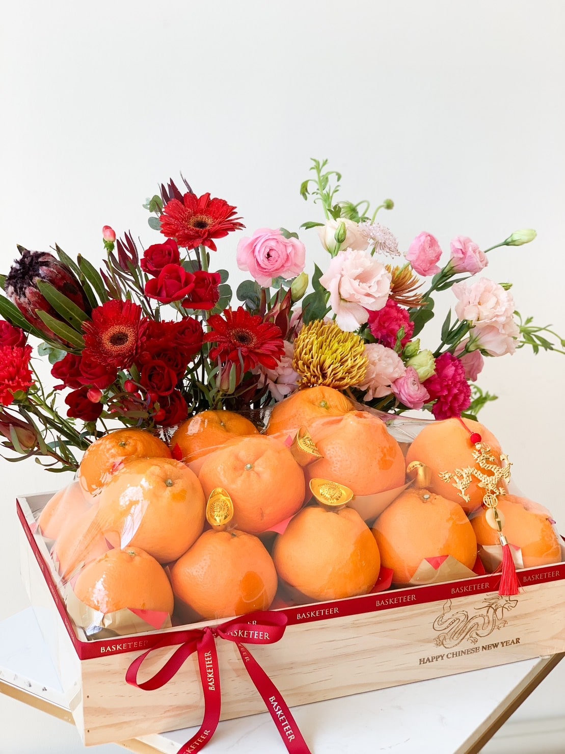 A wooden crate filled with vibrant oranges, adorned with red and gold ribbons, and decorated with a floral arrangement of red, pink, and yellow flowers. The crate is labeled 