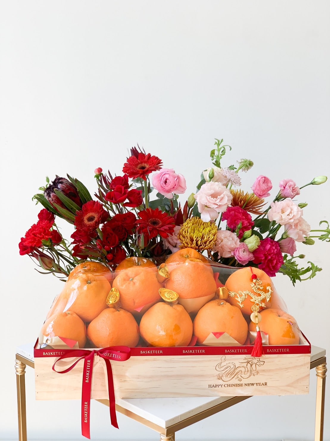 A wooden crate containing bright orange pomelos and adorned with a red ribbon sits on a table. Behind the pomelos, various vibrant flowers, including red, pink, and yellow blooms, are arranged. A tag reads “Happy Chinese New Year” on the crate.