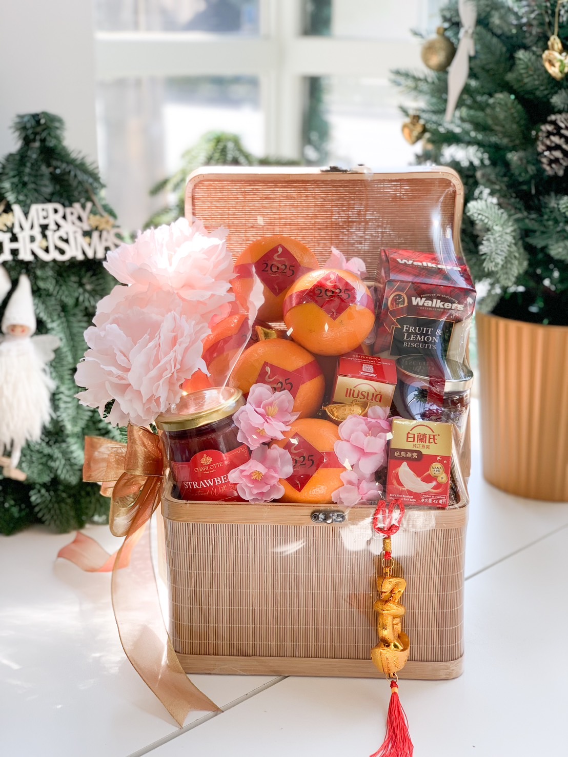 Chinese New Year gift basket in a woven case, filled with fresh oranges, strawberry preserves, festive treats, and adorned with pink flowers and gold-red ribbons.
