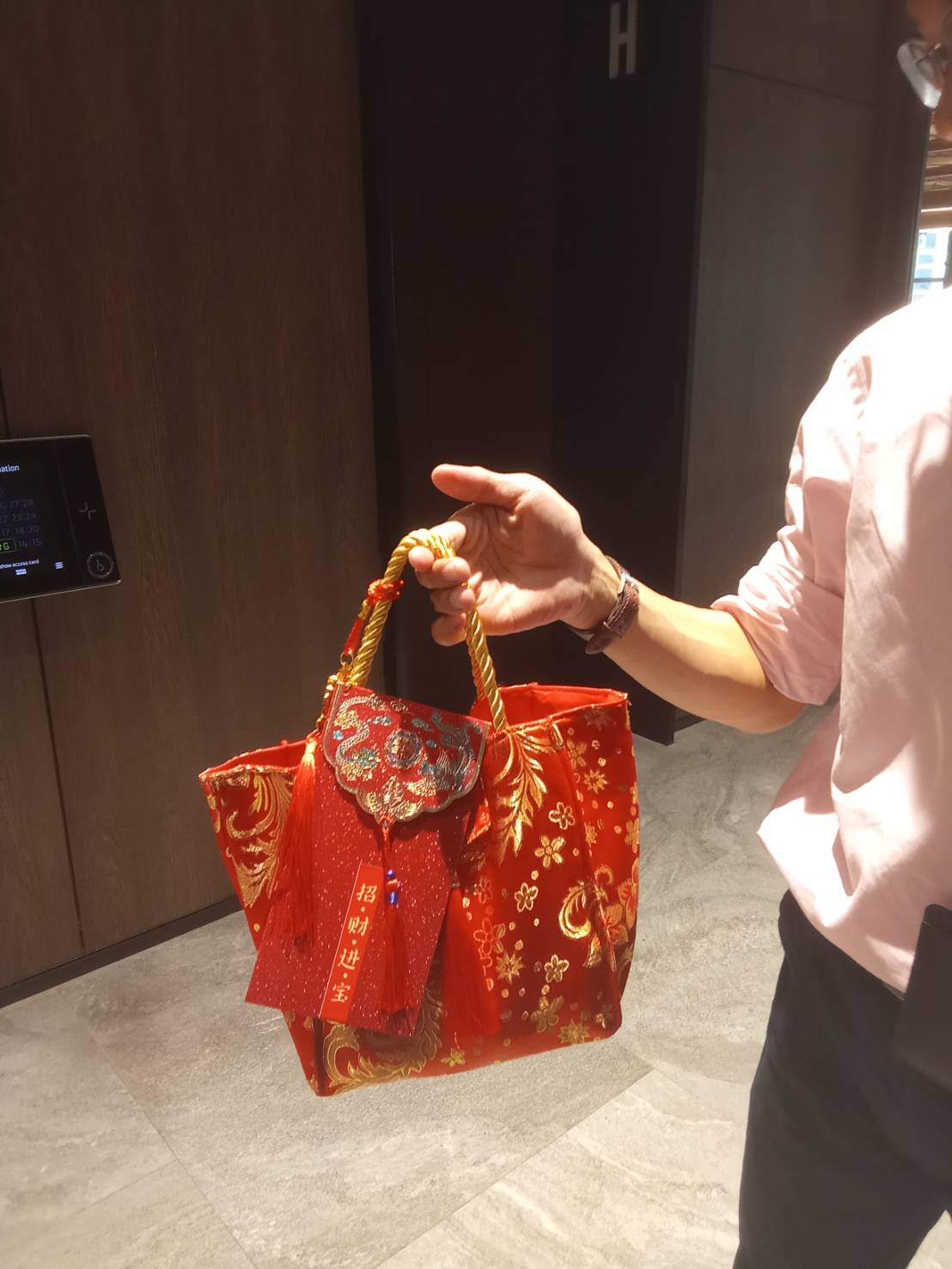 A person in a light pink shirt holds a vibrant red fabric handbag with intricate gold and silver patterns, reminiscent of a CNY Citrus Gift Bag. The bag features ornate decorations, including a detailed floral design and a twisted gold handle, topped off with a red tassel.