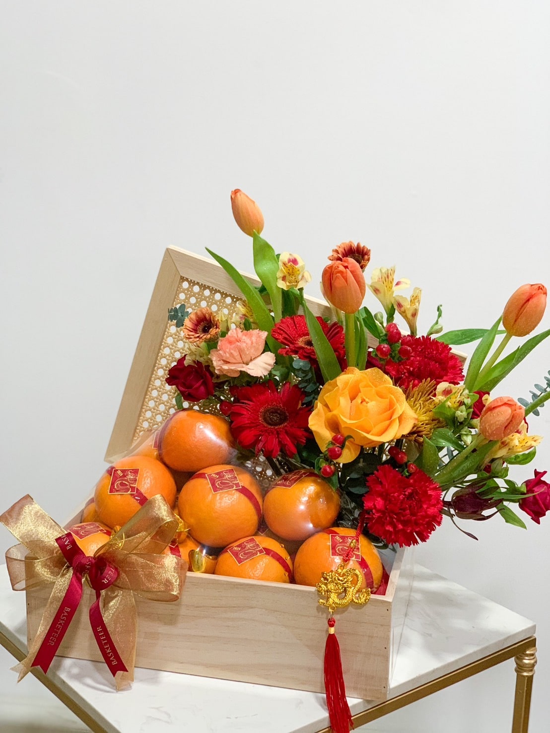 A wooden gift box filled with assorted fresh oranges is adorned with vibrant flowers including red roses, orange tulips, and yellow roses. A small red and gold gift bow is attached to a bunch of the oranges, and a decorative red tassel hangs from the box.