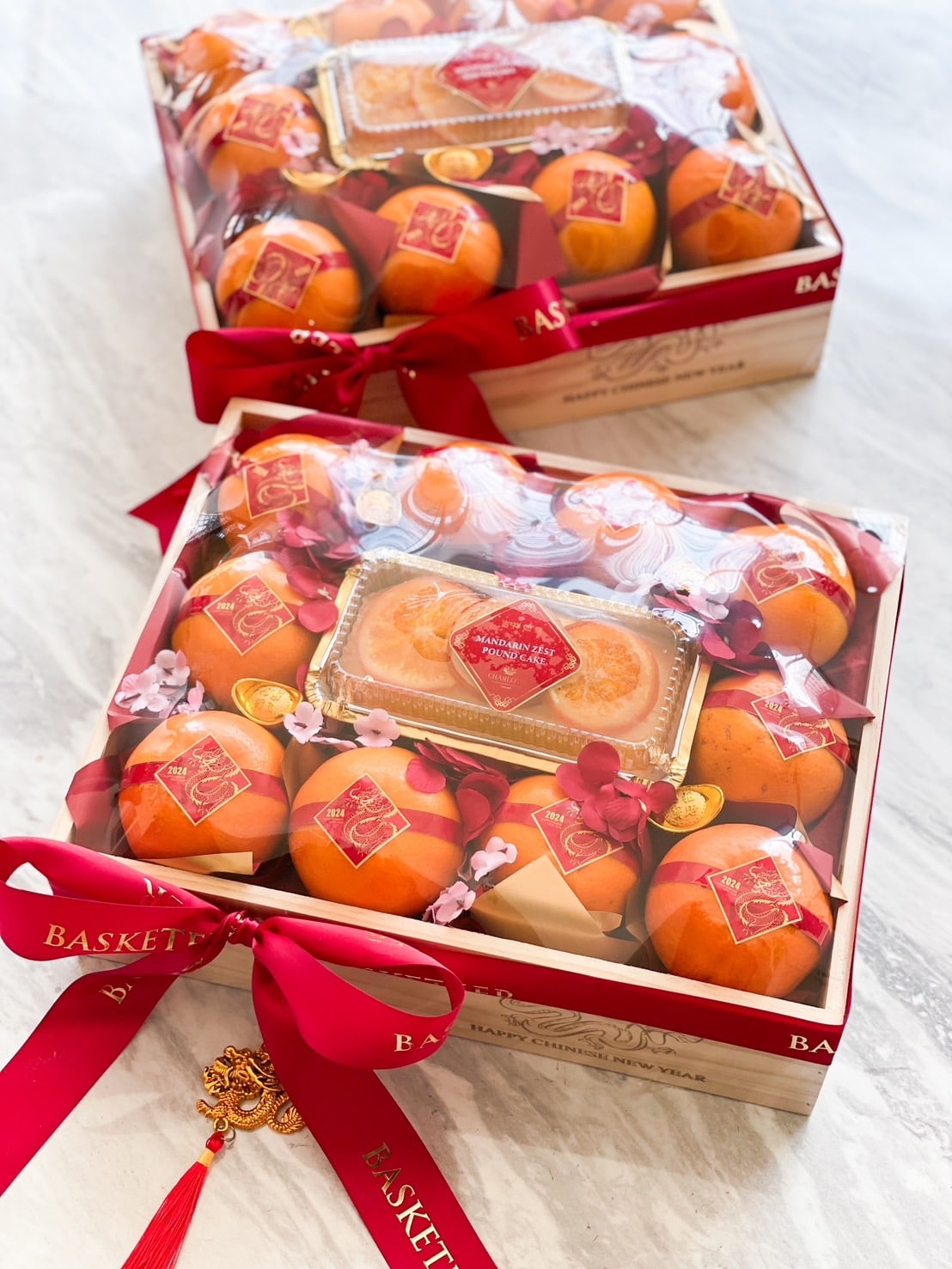 Two gift boxes containing fresh oranges and other fruits, wrapped in clear plastic and adorned with a red ribbon and a decorative charm. The boxes feature gold and red labels and are arranged on a light-colored marble surface.