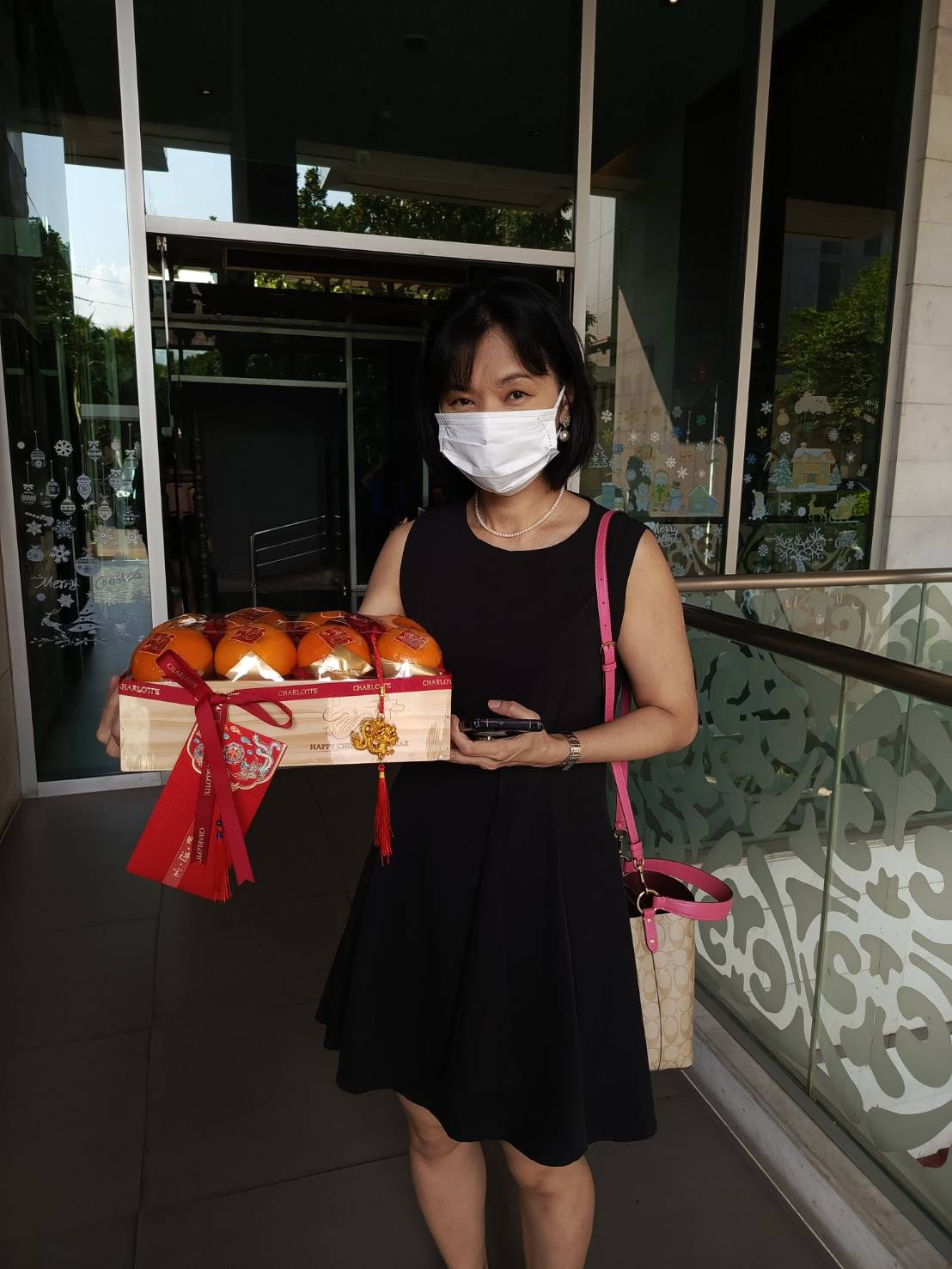 A woman in a black dress and white face mask stands in front of a building holding a Harmony Mandarin Woodcraft Delight crate filled with oranges. Adorned with red ribbons and a decorative ornament, the crate captures the essence of CNY. She has a pink handbag and a smartphone in her other hand.