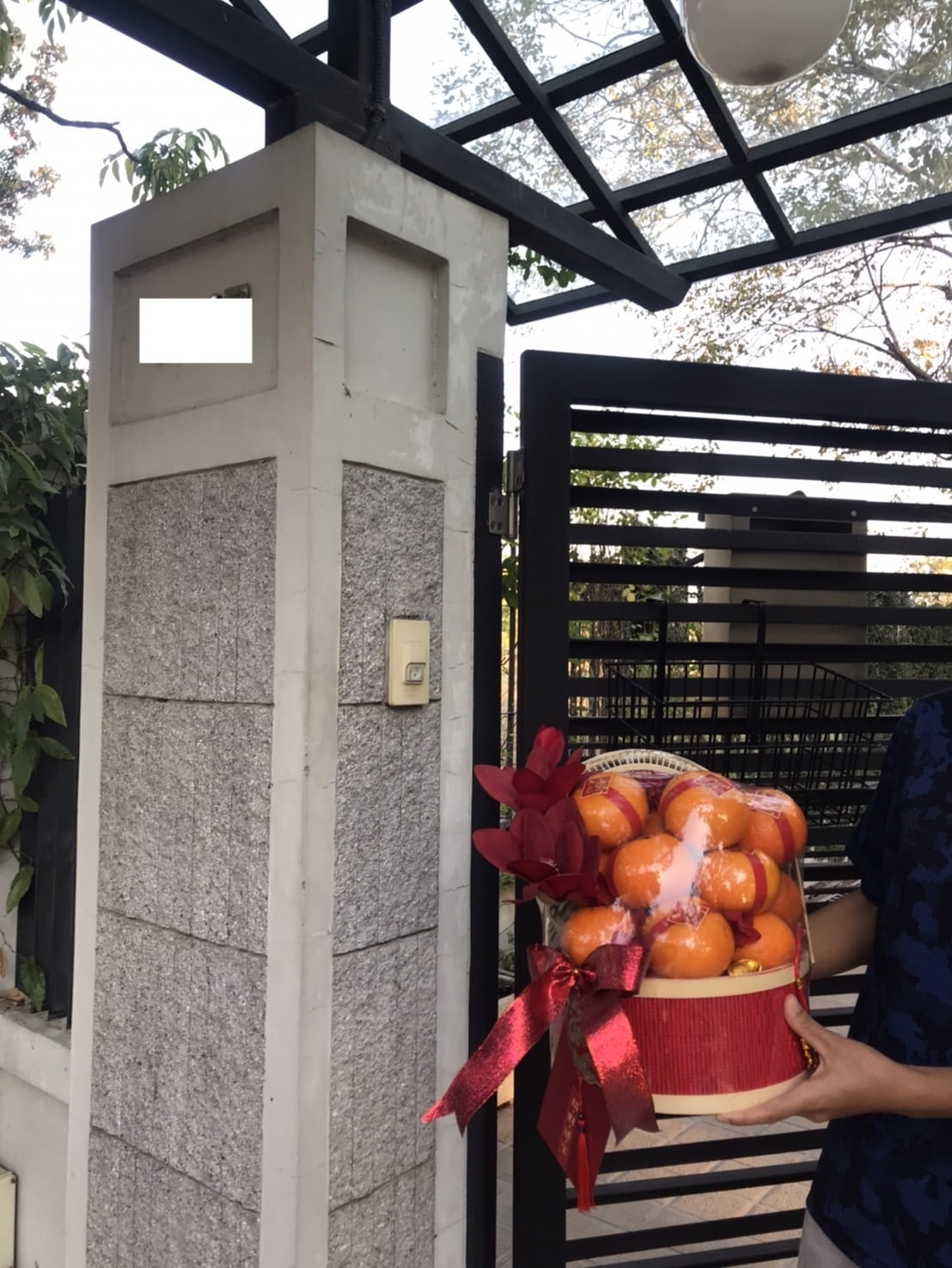 A person holding a CNY orange gift basket filled with oranges and wrapped with a red ribbon stands near the entrance gate of a house. The gate is partially open, revealing a stone pillar and some greenery in the background.